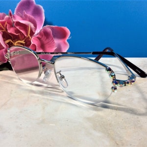 May include: Silver-framed reading glasses with clear lenses and iridescent crystal accents on the outer edges. The glasses have a half-rim design and black temples. The text "R376 +2.50" is printed on the frame.