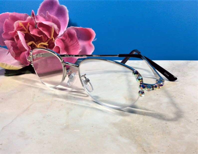 May include: Silver-framed reading glasses with clear lenses and iridescent crystal accents on the outer edges. The glasses have a half-rim design and black temples. The text "R376 +2.50" is printed on the frame.