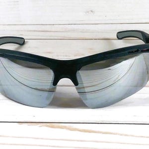 May include: Sport sunglasses featuring a matte black frame and reflective silver mirrored lenses. The design includes a wrap-around style with ventilated side temples and a contoured bridge, providing a wide field of vision for outdoor activities.