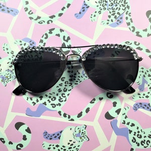 Pu&ograve; includere: Occhiali da sole aviator neri con un bordo di strass intorno alle lenti. Gli occhiali sono su uno sfondo rosa e verde con una stampa leopardata.