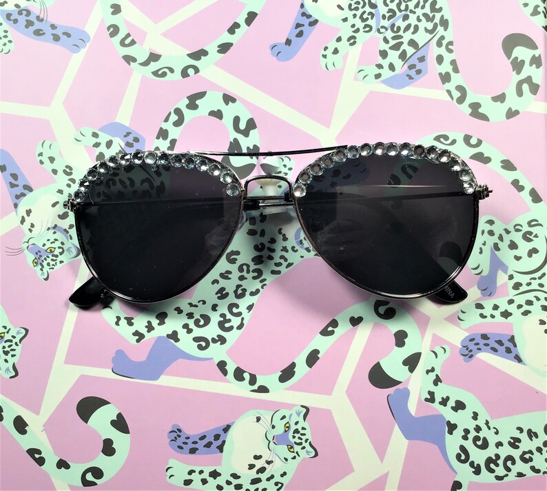 Pu&ograve; includere: Occhiali da sole aviator neri con un bordo di strass intorno alle lenti. Gli occhiali sono su uno sfondo rosa e verde con una stampa leopardata.