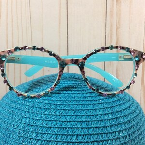 May include: A pair of eyeglasses with a light blue frame and temples. The rims are decorated with teal and purple rhinestones. The lenses are clear. The glasses are resting on a blue hat.