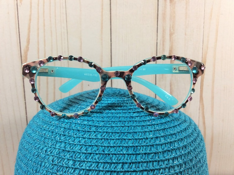 May include: A pair of eyeglasses with a light blue frame and temples. The rims are decorated with teal and purple rhinestones. The lenses are clear. The glasses are resting on a blue hat.