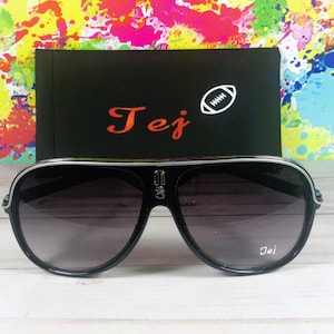 Puede incluir: Gafas de sol aviador negras con borde plateado y lentes degradadas. Las gafas de sol est&aacute;n frente a un estuche negro con el nombre "Tej" en escritura roja y un gr&aacute;fico de f&uacute;tbol americano blanco.