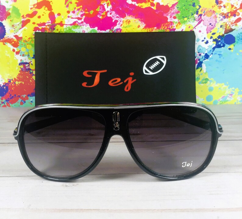 Puede incluir: Gafas de sol aviador negras con borde plateado y lentes degradadas. Las gafas de sol est&aacute;n frente a un estuche negro con el nombre "Tej" en escritura roja y un gr&aacute;fico de f&uacute;tbol americano blanco.
