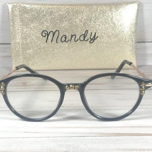 May include: Black-rimmed eyeglasses featuring gold-toned metal temples and small gold studs on the front frame corners and bridge. Behind the glasses sits a rectangular gold glitter case with the name Mandy written in black cursive script.