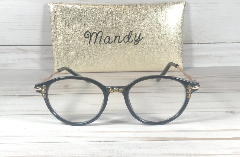 May include: Black-rimmed eyeglasses featuring gold-toned metal temples and small gold studs on the front frame corners and bridge. Behind the glasses sits a rectangular gold glitter case with the name Mandy written in black cursive script.