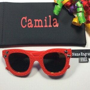 Puede incluir: Gafas de sol rojas con detalles de estrellas negras y una funda negra con el nombre "Camila" bordado. La funda est&aacute; junto a una etiqueta que dice "Nombre Grabado GRATIS".