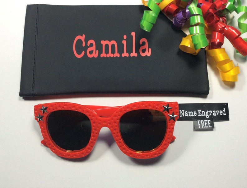 Puede incluir: Gafas de sol rojas con detalles de estrellas negras y una funda negra con el nombre "Camila" bordado. La funda est&aacute; junto a una etiqueta que dice "Nombre Grabado GRATIS".