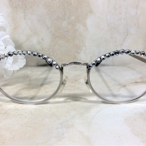 May include: Silver-rimmed eyeglasses with clear lenses. The top of the frames are embellished with a row of sparkling, clear crystals. The glasses have a delicate, elegant design, suitable for fashion or everyday wear.