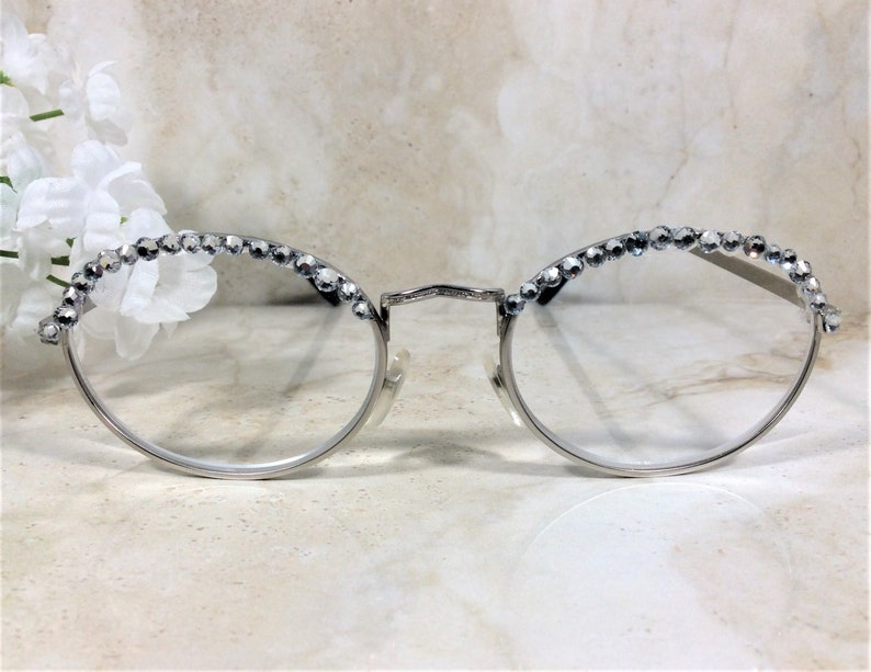 May include: Silver-rimmed eyeglasses with clear lenses. The top of the frames are embellished with a row of sparkling, clear crystals. The glasses have a delicate, elegant design, suitable for fashion or everyday wear.