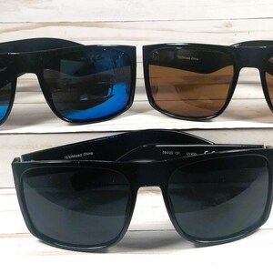 Puede incluir: Se muestran cuatro pares de gafas de sol con montura negra. Dos pares tienen lentes marrones, uno tiene lentes con espejo azul y otro tiene lentes grises oscuros. Las gafas de sol est&aacute;n sobre una superficie blanca.