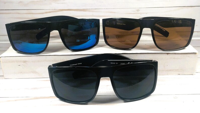 Puede incluir: Se muestran cuatro pares de gafas de sol con montura negra. Dos pares tienen lentes marrones, uno tiene lentes con espejo azul y otro tiene lentes grises oscuros. Las gafas de sol est&aacute;n sobre una superficie blanca.