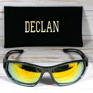 Może przedstawiać: Czarne okulary przeciwsłoneczne ze złotymi lustrzanymi szkłami i czarnym etui z napisem "DECLAN" nadrukowanym złotymi literami.