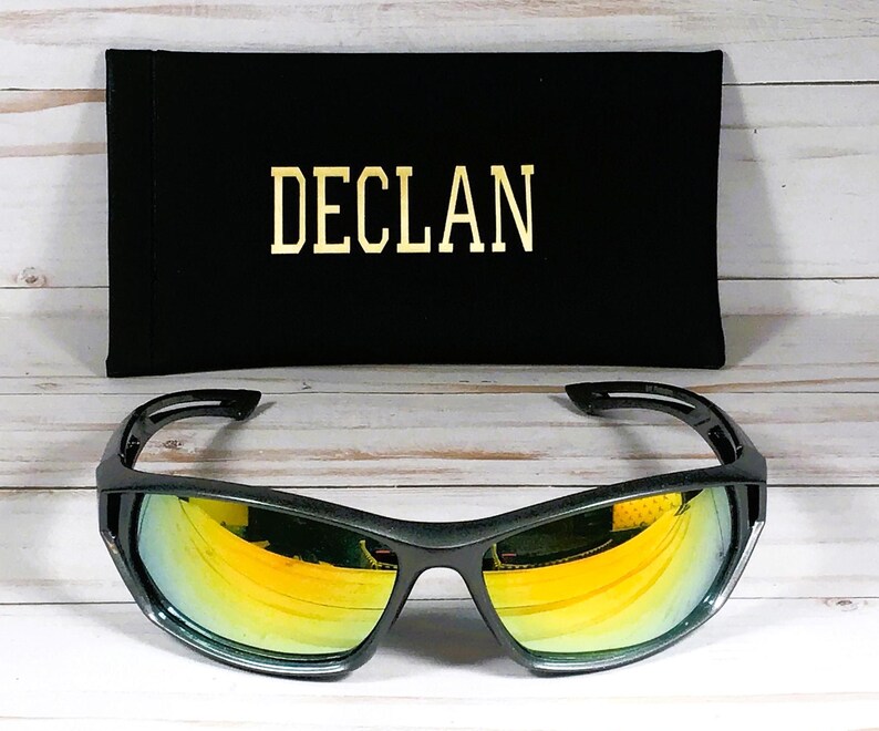 Może przedstawiać: Czarne okulary przeciwsłoneczne ze złotymi lustrzanymi szkłami i czarnym etui z napisem "DECLAN" nadrukowanym złotymi literami.