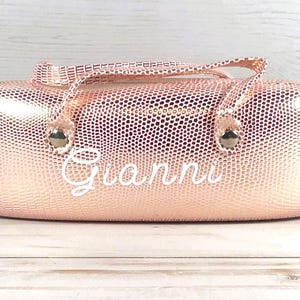 May include: A rose gold, textured glasses case with a handle and the name "Gianni" in white script. The case has a zip closure and metal accents. The background is a light wood surface.