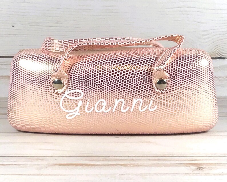 May include: A rose gold, textured glasses case with a handle and the name "Gianni" in white script. The case has a zip closure and metal accents. The background is a light wood surface.