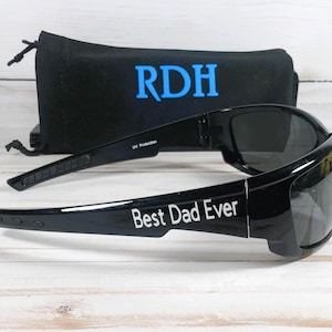 Puede incluir: Gafas de sol negras con lentes oscuras que presentan el texto blanco Best Dad Ever en la patilla. Detr&aacute;s de las gafas hay una bolsa de tela negra con cierre de cord&oacute;n, que muestra las letras azules RDH impresas en la parte frontal.