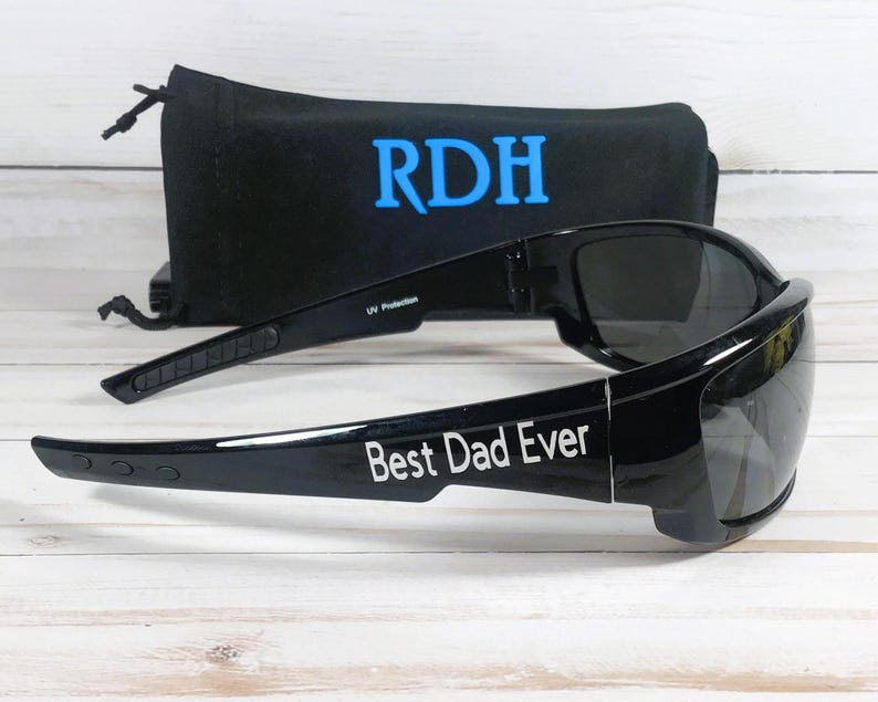Puede incluir: Gafas de sol negras con lentes oscuras que presentan el texto blanco Best Dad Ever en la patilla. Detr&aacute;s de las gafas hay una bolsa de tela negra con cierre de cord&oacute;n, que muestra las letras azules RDH impresas en la parte frontal.