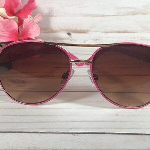 Pu&ograve; includere: Occhiali da sole aviator rosa con lenti marroni e dettagli dorati. Gli occhiali hanno una sottile montatura in metallo dorato e bordi in plastica rosa.