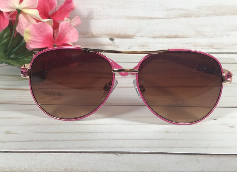 Pu&ograve; includere: Occhiali da sole aviator rosa con lenti marroni e dettagli dorati. Gli occhiali hanno una sottile montatura in metallo dorato e bordi in plastica rosa.