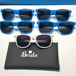 Puede incluir: Se muestran seis pares de gafas de sol. Cinco pares tienen monturas azul brillante y lentes oscuras. Un par tiene monturas blancas y lentes moradas, descansando sobre un estuche negro con la palabra "Bride" impresa.