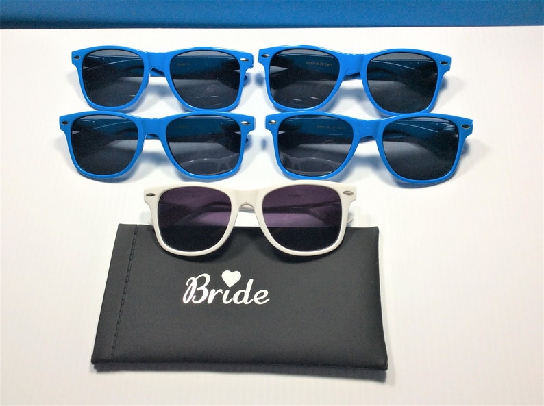 Puede incluir: Se muestran seis pares de gafas de sol. Cinco pares tienen monturas azul brillante y lentes oscuras. Un par tiene monturas blancas y lentes moradas, descansando sobre un estuche negro con la palabra "Bride" impresa.
