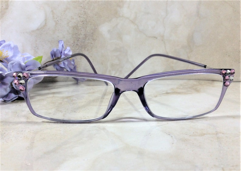 May include: Rectangular eyeglasses with a translucent purple frame. The outer corners of the frame are embellished with small, sparkling pink and clear rhinestones. The temples are thin and match the purple color of the front frame.