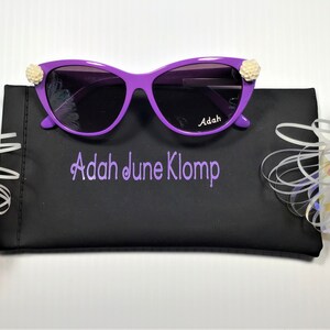 May include: Purple cat-eye sunglasses with white flower accents on the temples. The sunglasses are in a black case with the name "Adah June Klomp" embroidered on it.