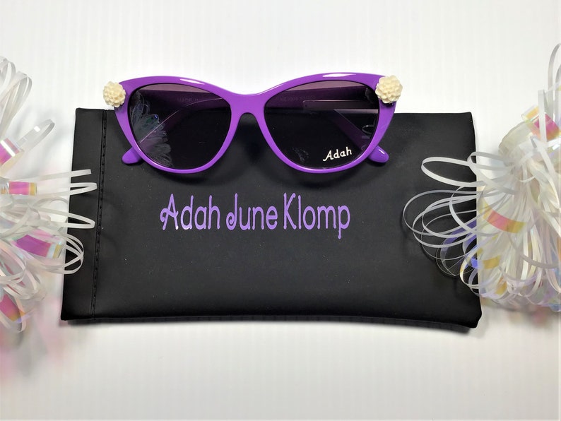 May include: Purple cat-eye sunglasses with white flower accents on the temples. The sunglasses are in a black case with the name "Adah June Klomp" embroidered on it.