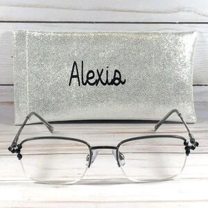 May include: A pair of eyeglasses with a silver frame and clear lenses, resting in front of a white and silver speckled case. The case has the name "Alexis" embroidered in black script.