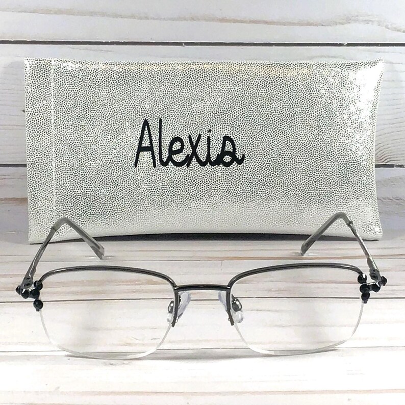 May include: A pair of eyeglasses with a silver frame and clear lenses, resting in front of a white and silver speckled case. The case has the name "Alexis" embroidered in black script.