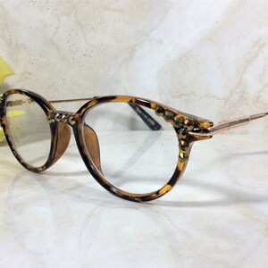 May include: Round eyeglasses with a tortoiseshell pattern frame and gold-colored metal arms. The frame is embellished with small, gold-colored studs. The lenses are clear. The glasses are set against a marble background.