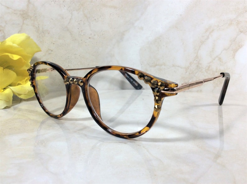 May include: Round eyeglasses with a tortoiseshell pattern frame and gold-colored metal arms. The frame is embellished with small, gold-colored studs. The lenses are clear. The glasses are set against a marble background.