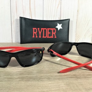 Op de afbeelding: Zwarte zonnebril met rode accenten en een gouden logo. De zonnebril wordt geleverd met een zwarte etui met het woord "RYDER" in rode letters en een witte ster.