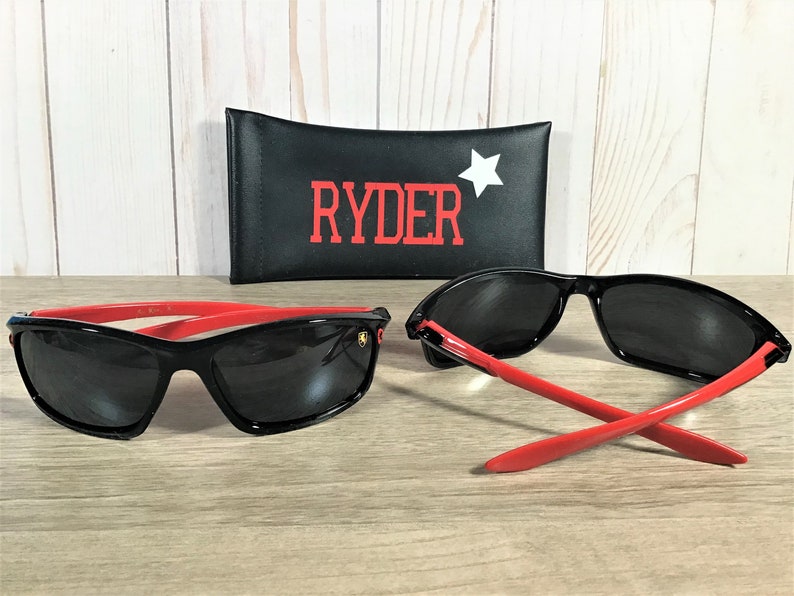 Op de afbeelding: Zwarte zonnebril met rode accenten en een gouden logo. De zonnebril wordt geleverd met een zwarte etui met het woord "RYDER" in rode letters en een witte ster.