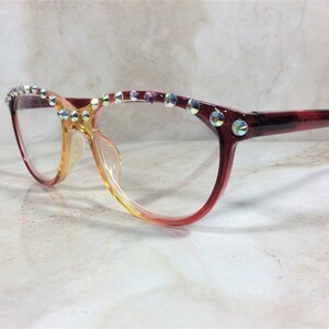 Puede incluir: Gafas con una montura degradada que pasa de un marr&oacute;n rojizo intenso en las varillas a un tono &aacute;mbar m&aacute;s claro en el puente. El borde superior de la montura est&aacute; adornado con una hilera de pedrer&iacute;a iridiscente de varios colores.