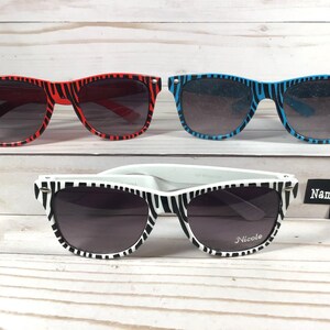 May include: Three pairs of zebra print sunglasses. The first pair is red, the second pair is blue, and the third pair is white with black stripes. The white pair has the name "Nicole" engraved on the right lens. The text "Name Engrave FREE" is visible in the image.