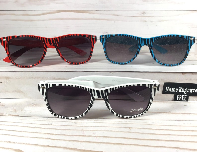 May include: Three pairs of zebra print sunglasses. The first pair is red, the second pair is blue, and the third pair is white with black stripes. The white pair has the name "Nicole" engraved on the right lens. The text "Name Engrave FREE" is visible in the image.