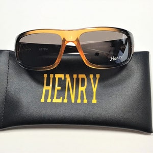 May include: Brown and orange sunglasses with a black case. The case has the word "HENRY" printed on it in gold letters. The sunglasses have the number "6777M" printed on the inside of the right lens.
