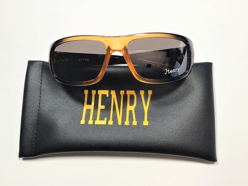 May include: Brown and orange sunglasses with a black case. The case has the word "HENRY" printed on it in gold letters. The sunglasses have the number "6777M" printed on the inside of the right lens.