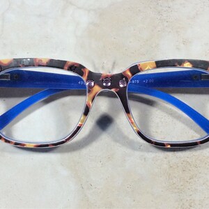 May include: Tortoiseshell-patterned reading glasses with blue arms and a square frame. The glasses have pink rhinestones on the top corners and the text "+2.00" is visible on the lens.