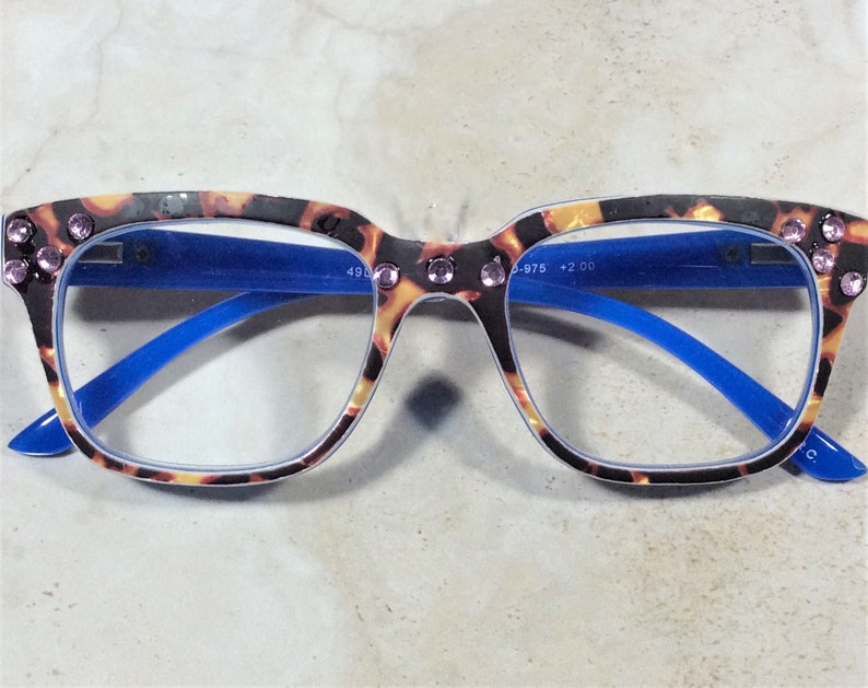 May include: Tortoiseshell-patterned reading glasses with blue arms and a square frame. The glasses have pink rhinestones on the top corners and the text "+2.00" is visible on the lens.