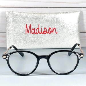 May include: Black-rimmed spectacles with clear lenses and small clear crystals embedded on the outer corners of the frames. The temples are thin and metallic gold. Behind the glasses is a rectangular silver glittery case with the name Madison in red.
