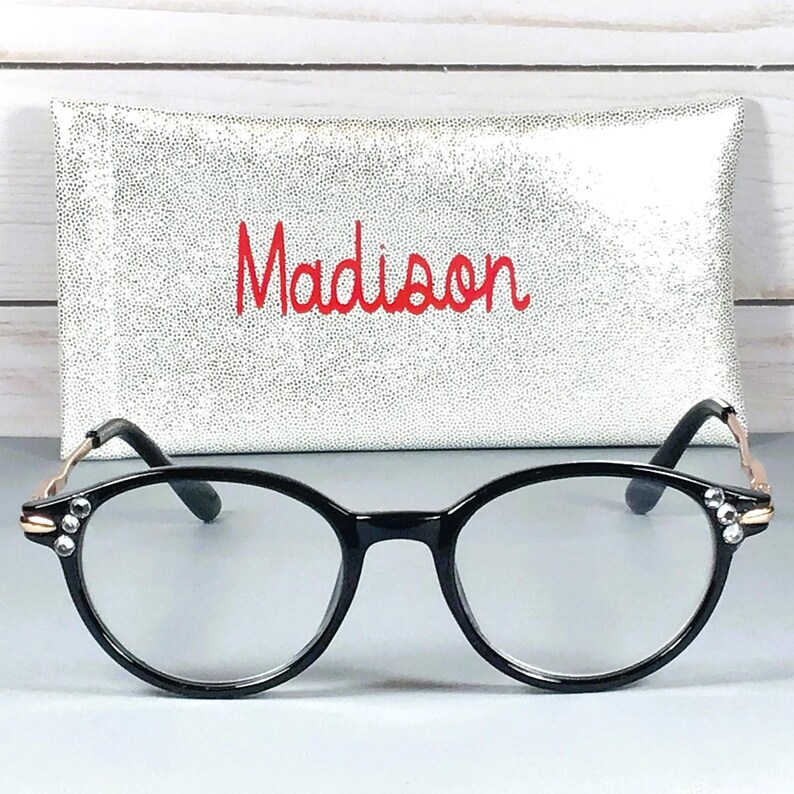 May include: Black-rimmed spectacles with clear lenses and small clear crystals embedded on the outer corners of the frames. The temples are thin and metallic gold. Behind the glasses is a rectangular silver glittery case with the name Madison in red.