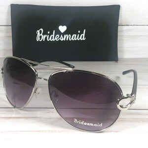 May include: Silver-framed aviator sunglasses with gradient purple lenses. The text "Bridesmaid" is printed on the bottom of the lenses. A black case with the word "Bridesmaid" in white script is in the background.