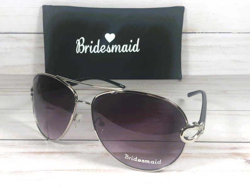 May include: Silver-framed aviator sunglasses with gradient purple lenses. The text "Bridesmaid" is printed on the bottom of the lenses. A black case with the word "Bridesmaid" in white script is in the background.