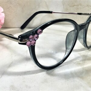 May include: Oval-shaped eyeglasses with a dark gray frame and clear lenses. The temples are silver-toned, and the frame is embellished with clusters of pink rhinestones. The glasses are set against a soft, neutral background.