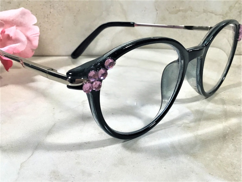 May include: Oval-shaped eyeglasses with a dark gray frame and clear lenses. The temples are silver-toned, and the frame is embellished with clusters of pink rhinestones. The glasses are set against a soft, neutral background.