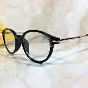 May include: Black eyeglasses with a round frame and gold-colored arms. The frames have decorative embellishments near the hinges. The lenses are clear. The glasses are set against a light-colored background.
